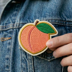 May include: Embroidered peach patch with a light orange fruit, a green leaf, and a brown stem. The patch has a cream-colored border and is being held against a denim jacket. The peach has a pink vertical line.