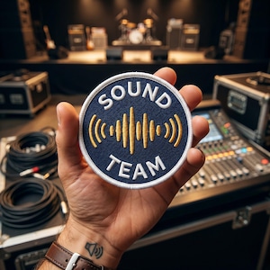 May include: A circular embroidered patch with a dark blue background, a white border, and white text reading SOUND TEAM. In the center, a gold-colored graphic depicts a vertical sound wave pattern. The patch is held against a blurred audio equipment background.