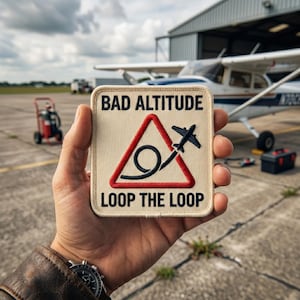 May include: A square patch with the text "BAD ALTITUDE" above a red triangle with a black airplane and loop-de-loop design. The text "LOOP THE LOOP" is below the triangle. The patch is held in a hand, with an airplane and hangar in the background.