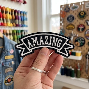May include: A black embroidered patch in the shape of a banner with white borders and the word AMAZING stitched in white capital letters across the center. The patch features a curved design with pointed ends on both the left and right sides.