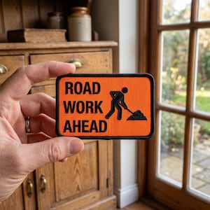 May include: An orange rectangular patch with black border and the words "ROAD WORK AHEAD" in white. A black silhouette of a worker shoveling dirt is on the right side of the patch. The patch is held up by a hand.