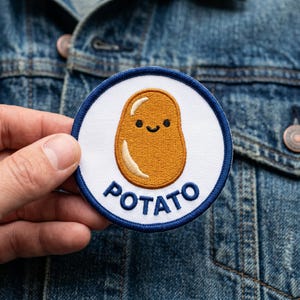 May include: Embroidered patch featuring a smiling cartoon potato in shades of brown and yellow, set against a white background with a navy blue border. The word "POTATO" is embroidered in navy blue below the potato.