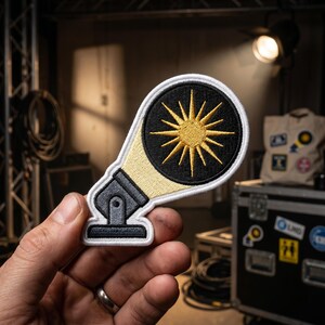 May include: An embroidered patch shaped like a stage spotlight. The base and neck are dark gray, while the light beam is light yellow. The circular lens features a black background with a bright yellow sunburst design, all outlined in white thread.