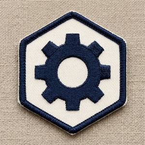 May include: Embroidered patch featuring a navy blue gear design on a white hexagonal background. The patch has a navy blue border and is set against a neutral fabric backdrop. The gear is a symbol of mechanics and engineering.