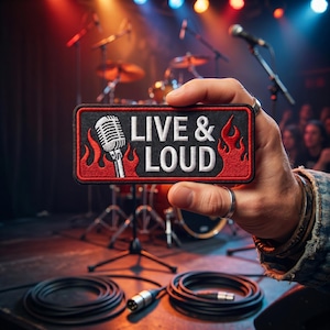 May include: A rectangular embroidered patch featuring a silver microphone icon on the left, red flames on the sides, and the white text "LIVE & LOUD" centered on a black background, held up against a blurred concert stage background.