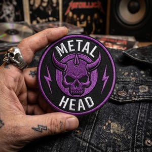 May include: A circular embroidered patch featuring a purple horned skull centered on a black background. The text METAL is arched above the skull and HEAD is arched below in white capital letters. Two purple lightning bolts flank the skull.