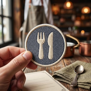 May include: A circular embroidered patch featuring a dark navy blue background with a light beige border. The center of the patch displays a light beige graphic icon of a fork and a knife positioned side by side in a vertical orientation.