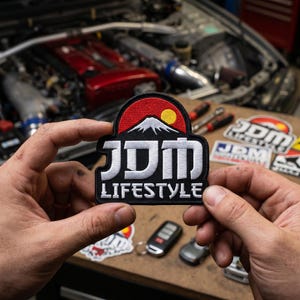 May include: A black and red patch with the text "JDM LIFESTYLE" in white. The patch features a mountain scene with a yellow sun. Other patches and car parts are visible in the background.