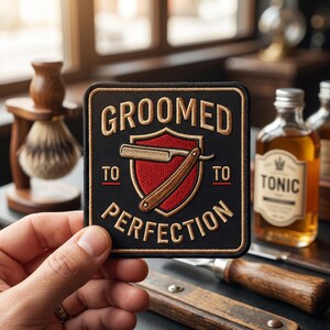 May include: A black and gold embroidered patch with the words "GROOMED TO PERFECTION" and a barber's straight razor design. The patch is held in a hand, with a blurred background of barber supplies.