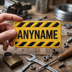 May include: A yellow and black patch with the word "ANYNAME" in bold black letters. The patch is rectangular with a black and yellow hazard stripe design. Tools and hardware are scattered on a wooden surface.
