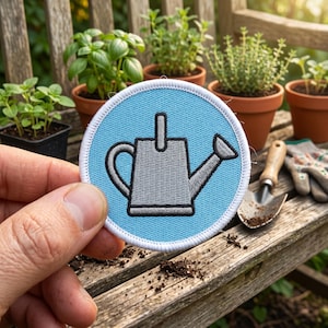 May include: A circular embroidered patch featuring a light blue background with a gray watering can icon in the center. The patch has a thick white stitched border. The design depicts a classic watering can with a handle and a spout.