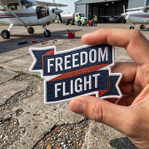 May include: A navy blue and white embroidered patch with the words "FREEDOM FLIGHT" in white block letters. The patch has a banner design with red and white accents. The patch is held in front of a small airplane.