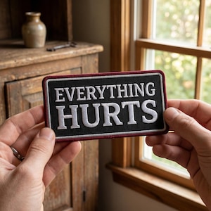 May include: A rectangular black patch with white embroidered text that reads "EVERYTHING HURTS." The patch has a burgundy border and is held between two hands. The background includes a wooden cabinet and a window.