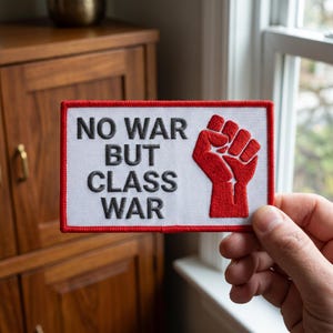 May include: A rectangular patch with a white background and a red border. The patch features the text "NO WAR BUT CLASS WAR" in black, with a red clenched fist graphic on the right side. The patch is held up by a hand.