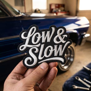 May include: A black and white embroidered patch with the words "Low & Slow" in a stylized script. The patch is held in front of a classic blue car in a garage setting.
