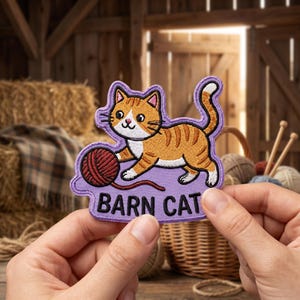 May include: An embroidered patch featuring an orange and white tabby cat playing with a red ball of yarn on a light purple background. The text BARN CAT is stitched in black capital letters at the bottom of the patch with a finished border.