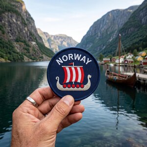 May include: A circular embroidered patch with a dark blue background featuring the white text NORWAY arched above a stylized beige Viking ship with a red and white striped sail and a row of red circular shields along the side of the hull.