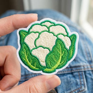 May include: Embroidered patch featuring a cauliflower design. The patch has a white cauliflower head with green leaves and a white border. The patch is held up by a person's fingers.