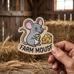May include: An embroidered patch featuring a gray mouse with pink inner ears holding a piece of yellow cheese, next to a larger wedge of Swiss cheese. The text FARM MOUSE is stitched in black at the bottom on a light beige background.