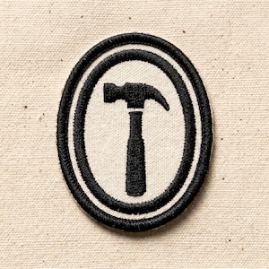 May include: Embroidered patch featuring a black hammer design within a white oval, framed by a double black border. The patch is set against a beige fabric background, suitable for sewing onto clothing or accessories.