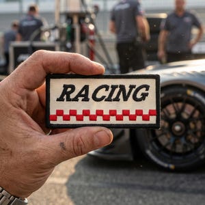 May include: A rectangular embroidered patch with a cream-colored background and a black border. The word RACING is stitched in bold black capital letters above a horizontal row of alternating red and white checkered squares across the bottom.