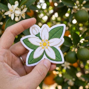 May include: An embroidered patch featuring a white citrus blossom with five petals, a yellow center, a small closed bud, and two green leaves. The design is outlined in white thread and features detailed stitching throughout the floral and leaf elements.