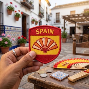 May include: A shield-shaped embroidered patch featuring a red border, a yellow center, and the word SPAIN in red capital letters. The patch displays a stylized orange and yellow folding fan graphic centered on the yellow background.