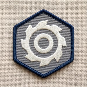 May include: Embroidered patch with a white gear design on a gray hexagonal background, framed by a navy blue border. The patch is likely for clothing or accessories.