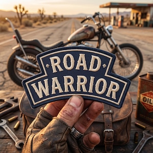 May include: A navy blue and tan embroidered patch with the words "ROAD WARRIOR" in a stylized font. The patch is held in front of a motorcycle, leather bag, and tools, with a desert highway in the background.