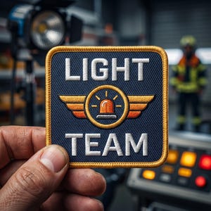 May include: A square embroidered patch with a dark blue background and a gold border. The text LIGHT TEAM is stitched in white. In the center, a circular emblem features orange wings and an orange emergency beacon light icon with rays.