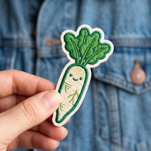 May include: Embroidered patch featuring a smiling white radish with green leafy top. The radish has a cute face and is outlined in green. The patch is held against a denim jacket.