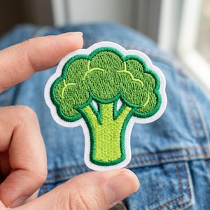 May include: Embroidered patch featuring a green broccoli design with a white border. The broccoli has a detailed texture and a bright green color, with a lighter green stem. This patch is ideal for adding to clothing or accessories.