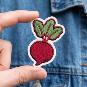 May include: Embroidered patch featuring a red beet with green leaves and red stems. The patch has a cream-colored border and is held against a denim jacket. The beet is a vibrant, detailed design.