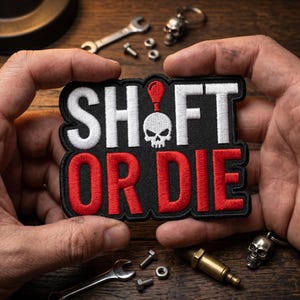 May include: Embroidered patch with the words "SHIFT OR DIE" in white and red on a black background. The patch features a skull graphic and is held by hands. Small tools and skull keychains are scattered around.
