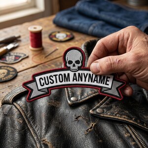 May include: A custom embroidered patch featuring a white skull icon centered above a grey banner with a red border. The banner contains the black text CUSTOM ANYNAME in a bold sans-serif font. The patch is designed for attachment to clothing.