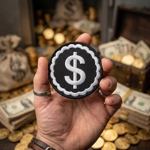 May include: A circular embroidered patch featuring a white dollar sign symbol centered on a black background, surrounded by a white scalloped border. The patch is held in a palm, with the design clearly visible against the dark fabric base.