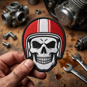 May include: Embroidered patch featuring a skull wearing a red and white racing helmet. The patch has a black border and is set against a wooden surface with motorcycle parts and tools.