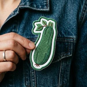 May include: A green embroidered patch in the shape of a zucchini, with a white border and green leaves. The patch is being held up against a denim jacket.