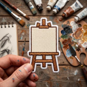May include: An embroidered patch shaped like an artist's easel with a brown wooden frame and a blank, light beige canvas center. The patch features a thick white border and detailed stitching that mimics the texture of wood grain on the easel legs.