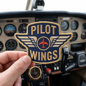 May include: A navy blue Pilot Wings patch with gold embroidered wings and lettering, featuring a red airplane in the center. The patch is held in front of an airplane dashboard.