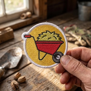May include: A circular embroidered patch featuring a yellow background with a white border. The design depicts a red wheelbarrow filled with a pile of green grass or hay, with a black wheel and a red handle, rendered in detailed thread work.