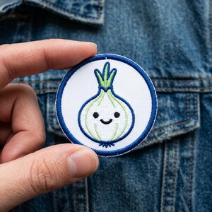 May include: A round embroidered patch featuring a smiling onion character. The onion is white and green, with a blue outline and border. The patch is held against a denim jacket.