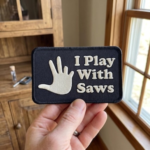 May include: A navy blue rectangular patch with the text "I Play With Saws" in white. A white hand gesture is on the left side of the patch. The patch is held by a person.
