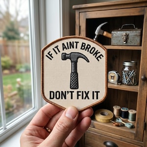 May include: A beige patch with the text "IF IT AINT BROKE DON'T FIX IT" and a black hammer graphic. The patch is held in a hand, with a wooden cabinet and window in the background.