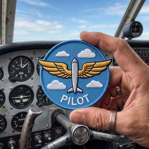 May include: A circular embroidered patch with a light blue background featuring a white airplane silhouette with gold wings, white clouds, and the word PILOT in white capital letters. The patch has a stitched border in a matching light blue thread.