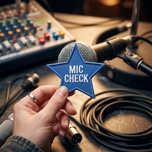 May include: A blue star-shaped embroidered patch with the white text MIC CHECK centered on the front. The patch features a textured fabric finish and is held in front of a silver metal microphone, audio cables, and a sound mixing console.