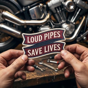 May include: A patch with the text "LOUD PIPES SAVE LIVES" in white lettering on a burgundy banner with a navy blue stripe. The patch is held in front of a motorcycle engine and exhaust pipes.