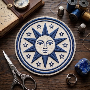 May include: Embroidered patch featuring a sun face with blue rays and stars on a cream background. The patch is surrounded by a blue border. Additional items include scissors, spools of thread, a compass, and a book on a wooden surface.