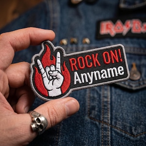 May include: A rectangular embroidered patch with a black background and a grey border. It features a white hand making a rock-and-roll sign in front of red flames. The text ROCK ON! is in red, and the word Anyname is in white below it.