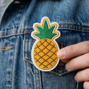 May include: Embroidered pineapple patch with a white border. The patch features a yellow body with brown crosshatch detailing and green leaves. The patch is being held against a denim jacket.